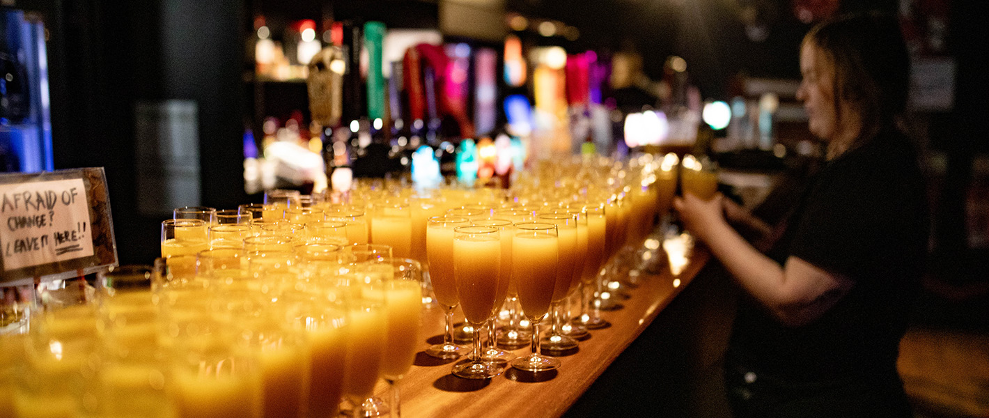 A bar covered in glasses of Mimosa cocktails with a white woman pouring more.