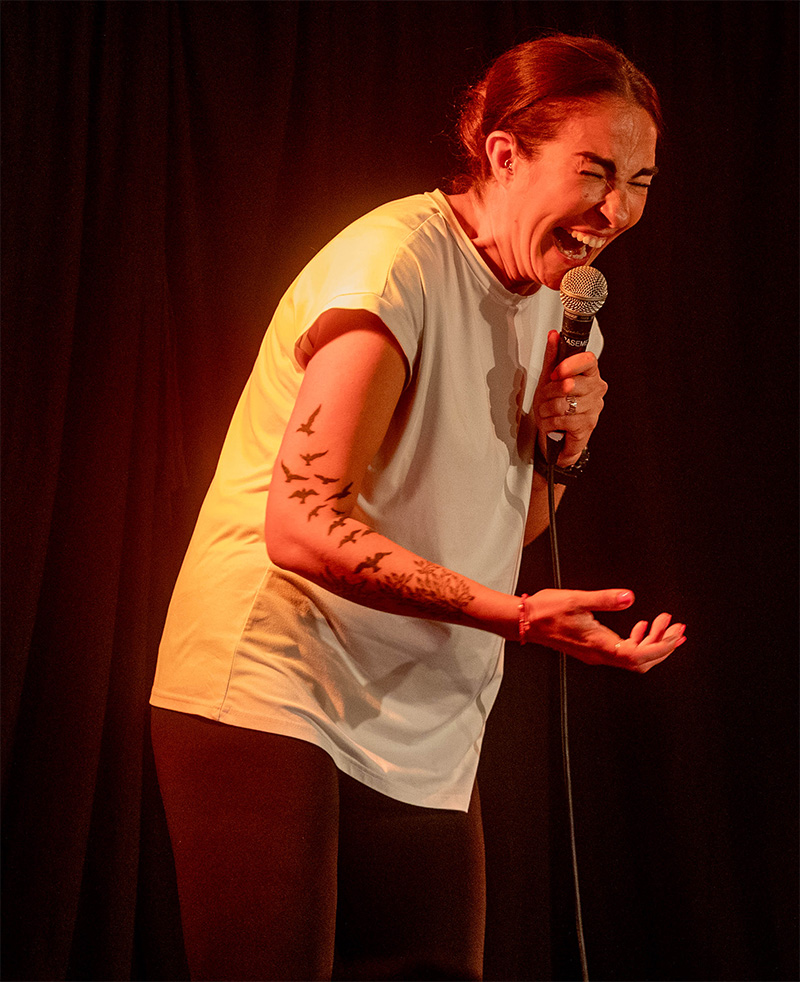 Esther Manito, a white woman with brown hair wearing a white t-shirt and black leggings, laughing into a microphone.