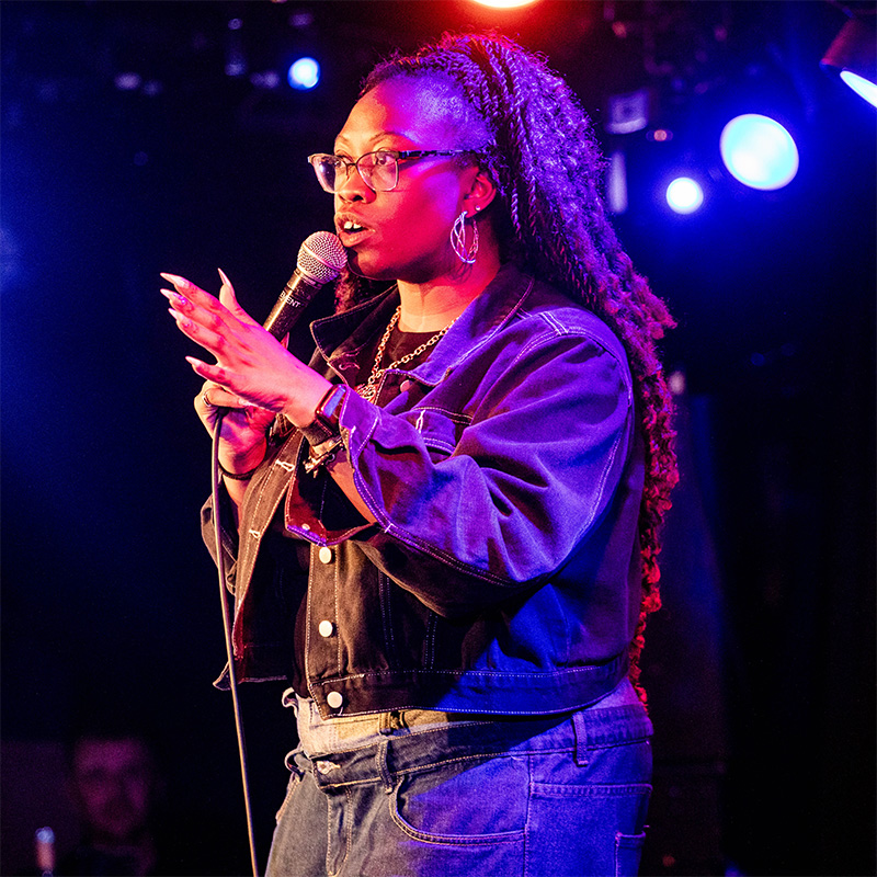 An image of Sikisa, a black woman wearing a denim jacket and jeans, speaking into a microphone on stage.