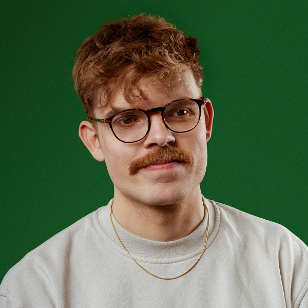 Horatio Gould, a white man with blonde hair, a moustache, black glasses and a beige sweatshirt on a green background.