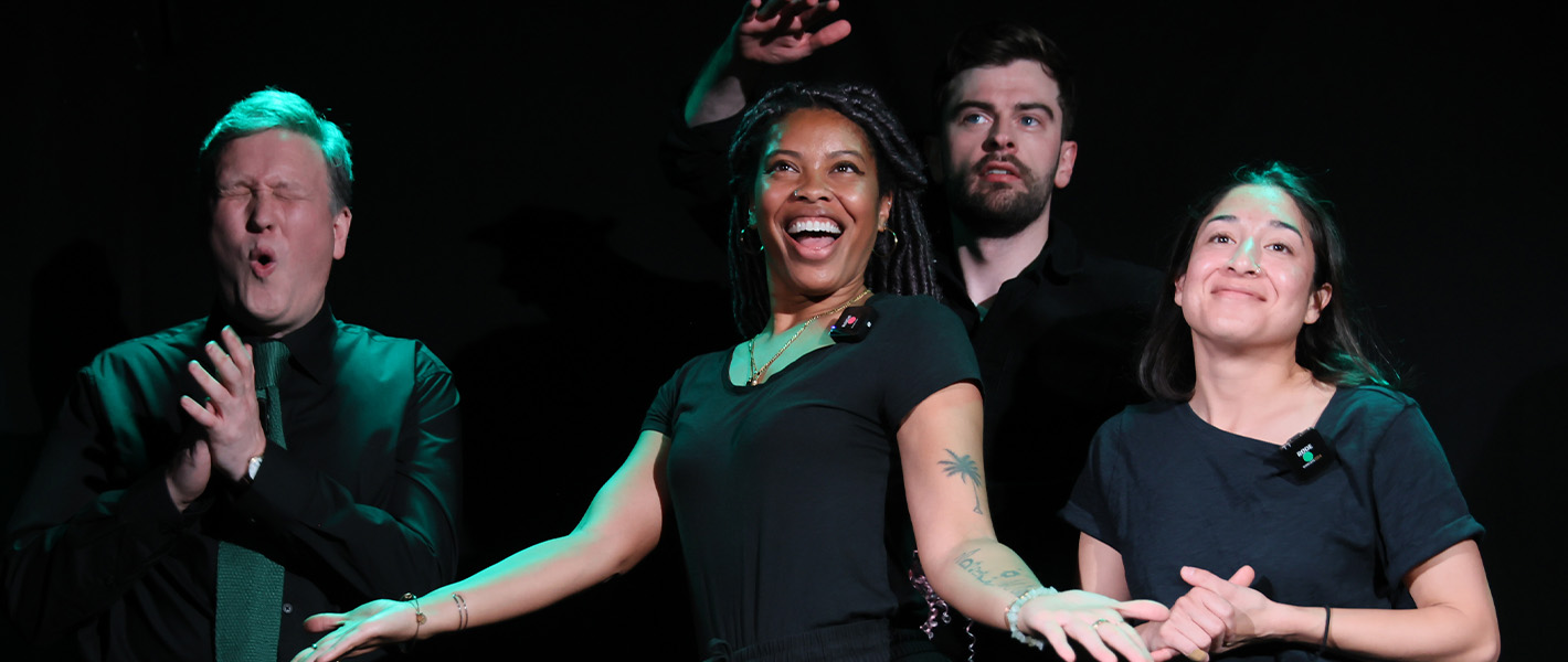 The Improbotics team on stage - left to right - a white men with blonde hair and a black shirt, a black woman with black braid and a black t-shirt, a white man with brown hair and a black shirt, a white woman with dark brown hair and a black shirt. On a black background.