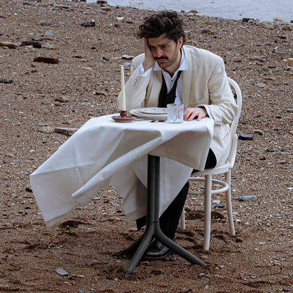 Paddy Young, a white man with dark brown hair and moustache, wearing a white dinner suit and undone bowtie, sat at a restaurant table on the beach, looking sad.